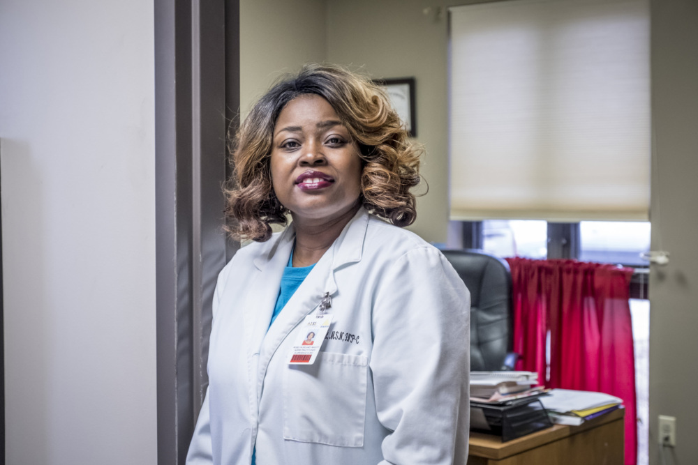 A person wearing a white lab coat stands in an office with a window in the background.